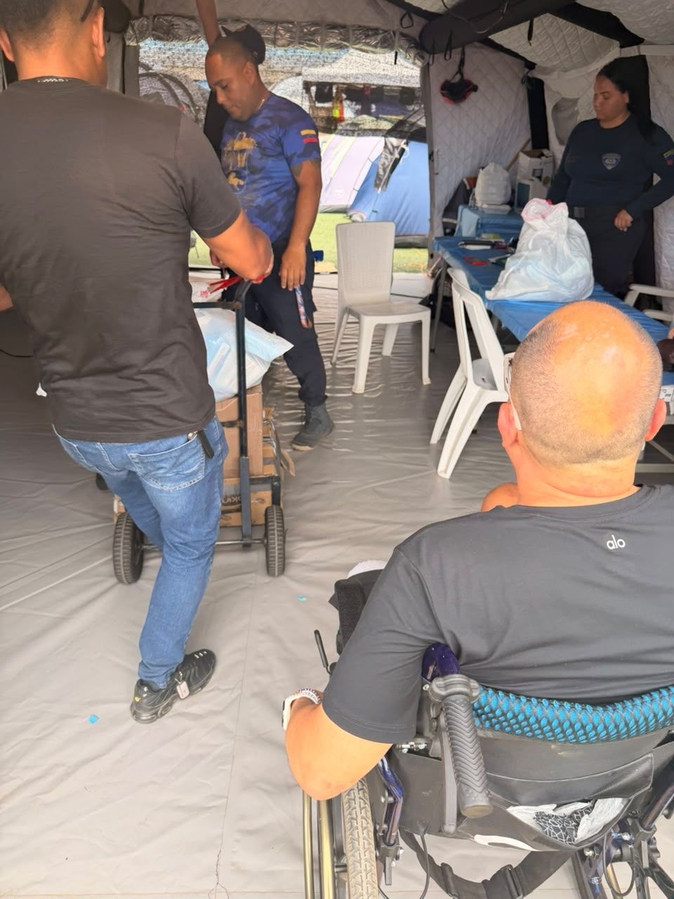 Un hombre empuja una carretilla con suministros hacia una carpa con mesas, observado por un hombre en silla de ruedas.